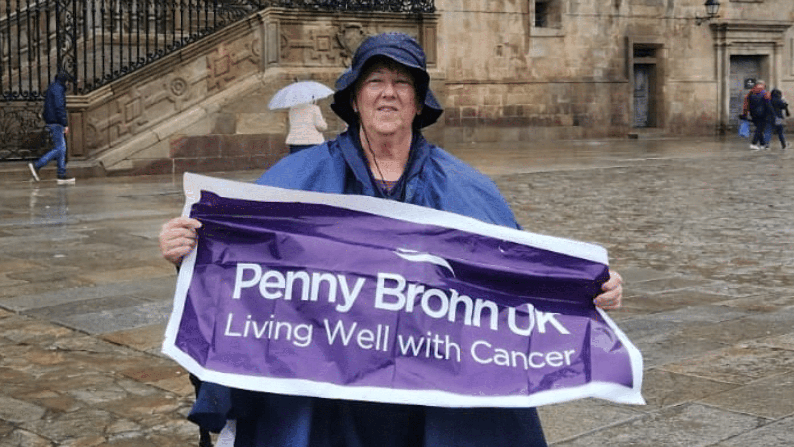 Photo of Heather Thomas wearing a hat and purple poncho holding a purple Penny Brohn UK banner.