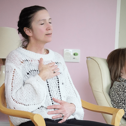 A woman sitting in an armchair with her hand on her chest and her eyes closed during a one-to-one therapy session.